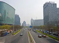 A wide divided street, seen from above, flanked by trees and medium-height high rises. The one on the left is somewhat curvilinear