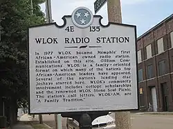 A white plaque with the designation 4E 135 at the top, reading "WLOK Radio Station: In 1977 WLOK became Memphis' first African-American owned radio station. Established on this site, WLOK is a family-oriented format on which many of the nation's top African-American leaders have appeared. Several of the nation's leading disc jockeys starred here. WLOK's community involvement includes college scholarships and the renowned WLOK Stone Soul Picnic. The station's call letters, WLOK-AM, are 'A Family Tradition.' "