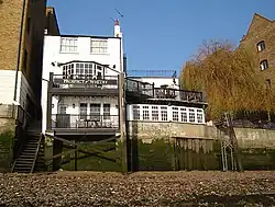 A historic riverside pub with multiple stories, traditional windows, and a dark exterior, situated on the bank of a wide river.