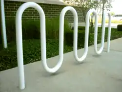 A white-painted round metal tube bent into a double M shape and set in concrete in front of a grassy verge and a brick wall.