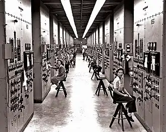 A long corridor with many consoles with dials and switches, attended by women seated on high stools.