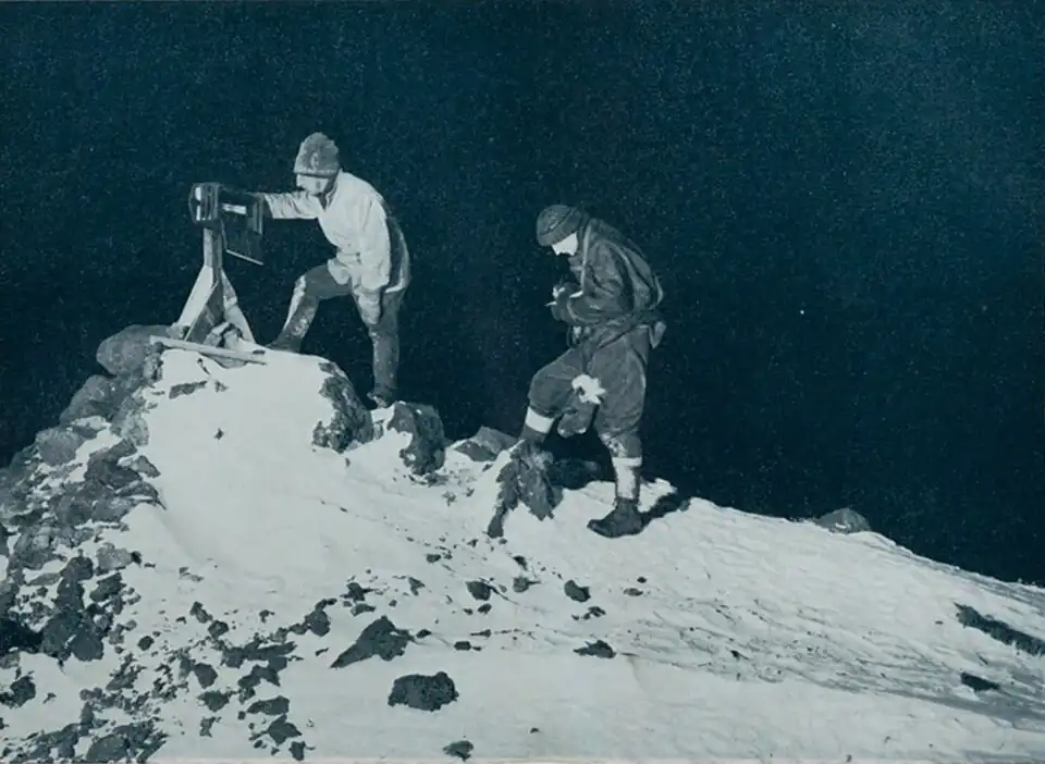 Photo of two men hunched by a box on a tripod at the top of a sharp hill