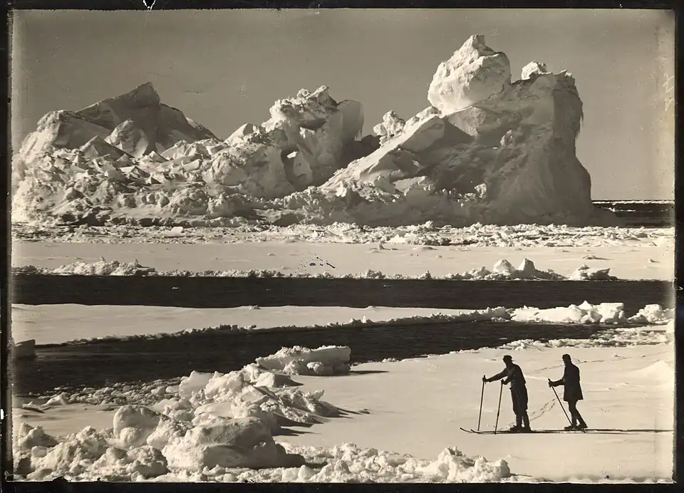 Photo of an eroded iceberg with two men skiing on an ice floe in the foreground