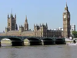 A colour photograph of the Palace of Westminster, containing the Houses of Parliament of the United Kingdom. Photograph shows a long neo-gothic building with a clock tower at the right, with Westminster Bridge and the River Thames in the foreground.