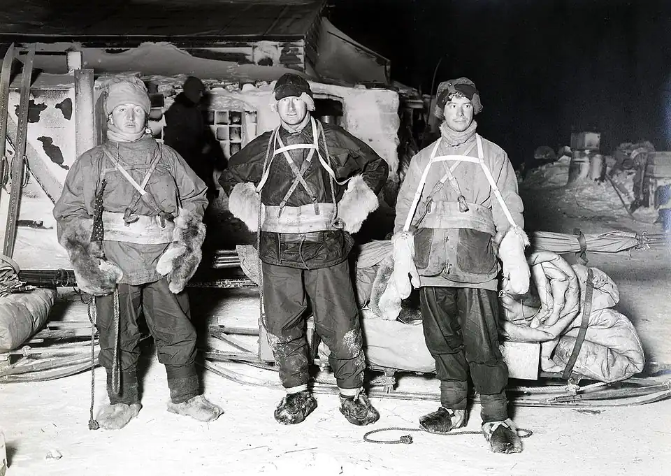 Photograph of three men in cold weather gear standing before a sled in polar darkness