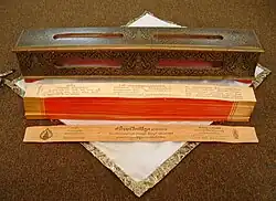 Colour photograph of a stack of long, thin wooden rectangles; each with scripture written on the face. The stack sits on a white cloth, in front of a carved wooden box.
