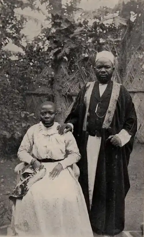 A man standing and a woman seated, both wearing formal attire