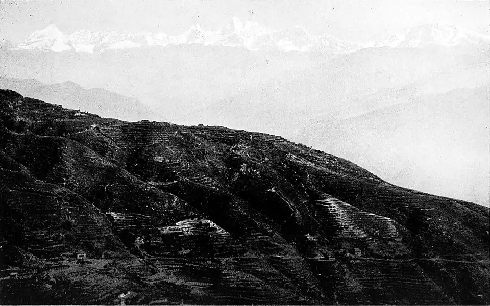 Black and white photograph of a mountain range. Half of the image is filled by the side of a mountain in the foreground, slanting down to the right. In the background, further faint mountain slopes can be seen. At the very top of these, at the furthest distance, are white, snow topped mountain peaks.