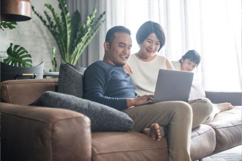 An Asian father and mother sit with their son on the couch looking at a laptop.